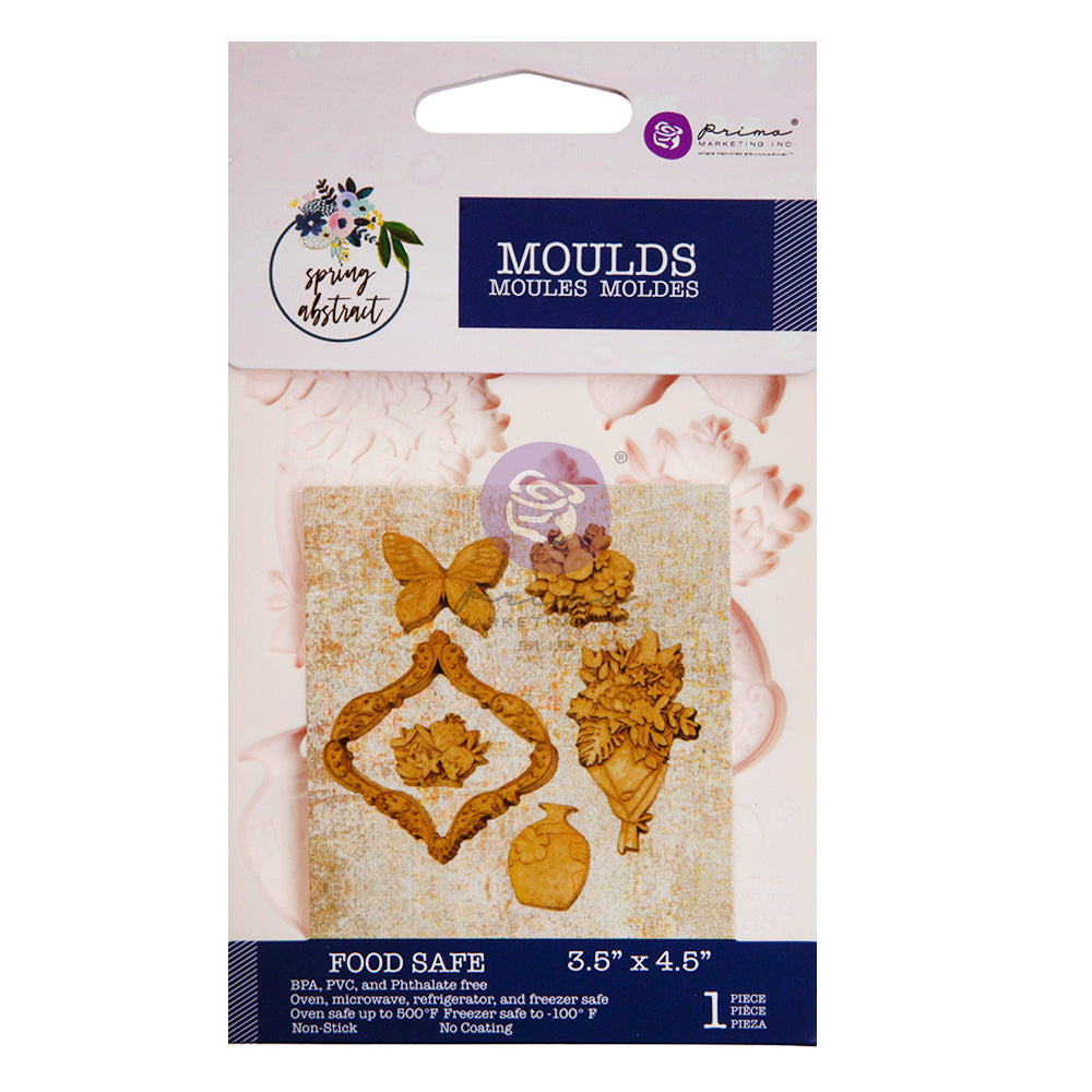 A retail package for a decorative silicone mould is displayed against a white background. The packaging is branded “Prima Marketing Inc.” and part of the “Spring Abstract” collection. The insert card shows sample embellishments in a natural wood-like finish, including a butterfly, floral clusters, a decorative ornate frame, a bouquet, and a small vase. The mould itself is pale pink and partially visible behind the insert. Text on the package indicates the mould is food safe and measures 3.5 by 4.5 inches. T