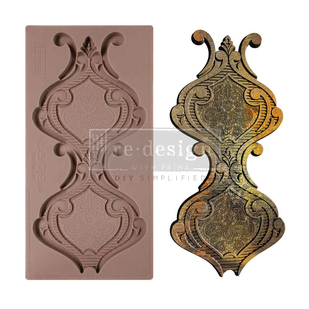 A side-by-side product display features a silicone décor mold on the left and a finished ornate casting on the right against a plain white background. The brown silicone mold contains two vertically stacked baroque-style plaque designs with symmetrical scrolling flourishes, pointed leaf accents, and finely textured ornamental centers. The completed casting on the right is painted in layered metallic tones of gold, bronze, and black, emphasizing the intricate filigree patterns and carved details. The elongat