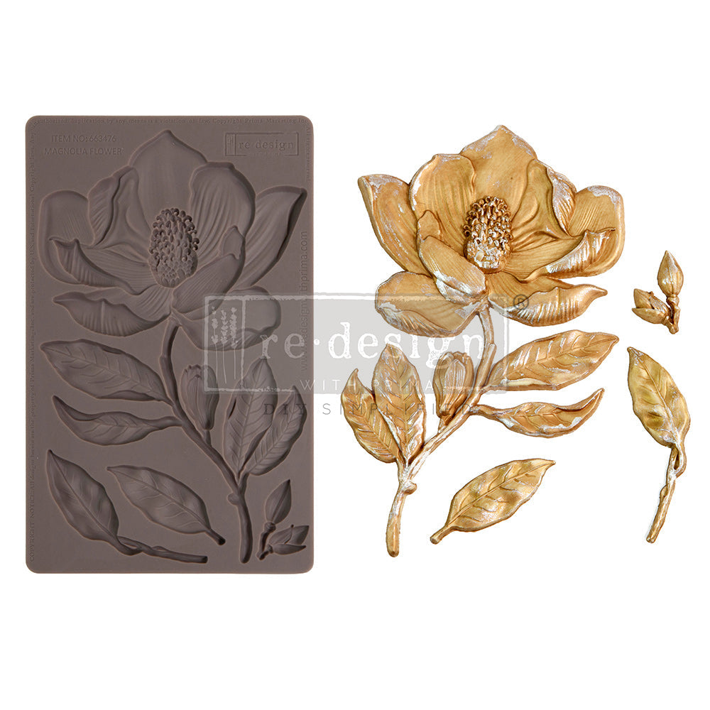 A product-style image displays a decorative silicone mould alongside completed floral cast embellishments. On the left is a rectangular silicone mould in a muted taupe-brown color featuring recessed impressions of a large magnolia flower with layered petals, branching stems, leaves, and a small bud. The mould captures detailed veining and botanical textures throughout the design.