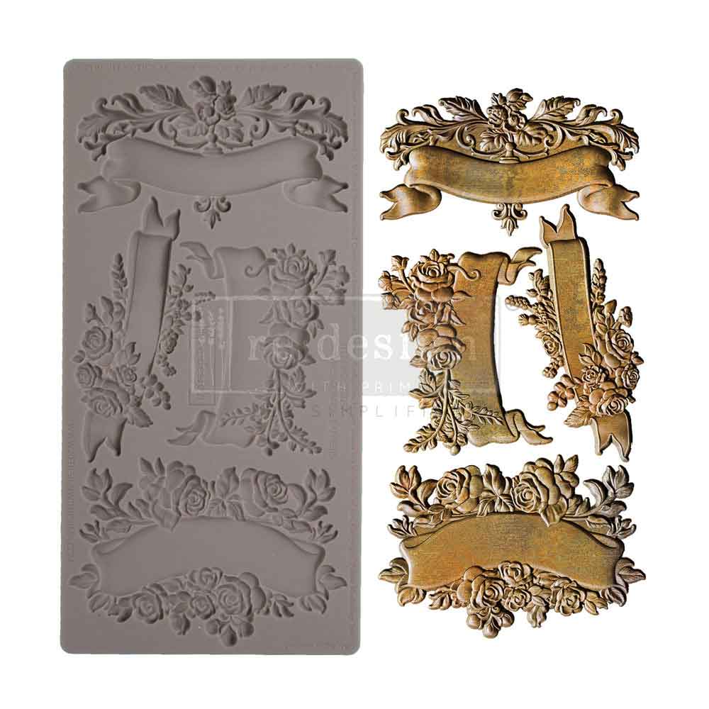 A product display showcases a silicone décor mould alongside finished cast embellishments in an antique gold finish. On the left is a tall rectangular gray silicone mould featuring recessed floral banner and scroll designs. On the right are the completed decorative pieces, including ornate ribbon banners framed with roses, leaves, and curling botanical details. The embellishments have a distressed metallic gold appearance with dark antiqued shading that highlights the intricate textures. The overall style i